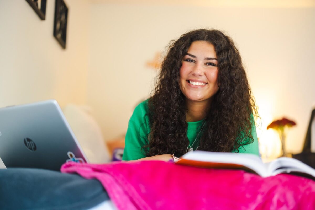 studing laying on a bed in a residence hall studying on a laptop with a book open