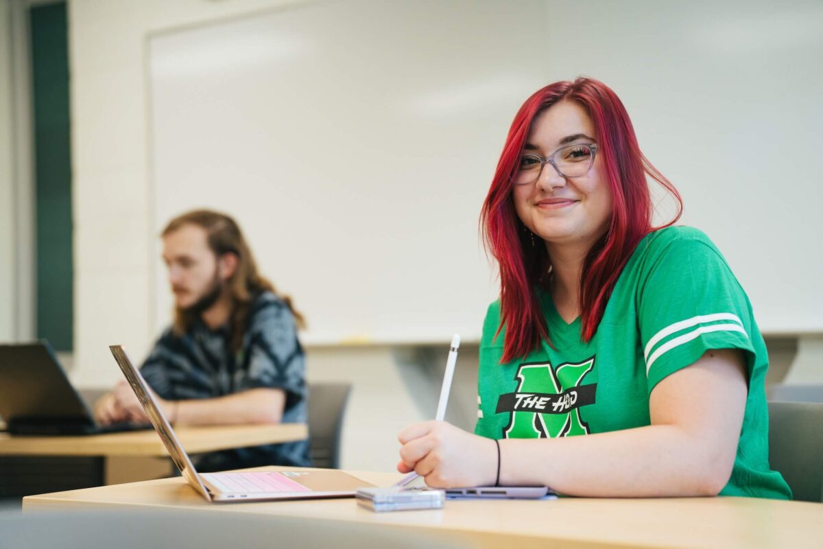 two students in class, one looking at the camera and smiling and taking notes