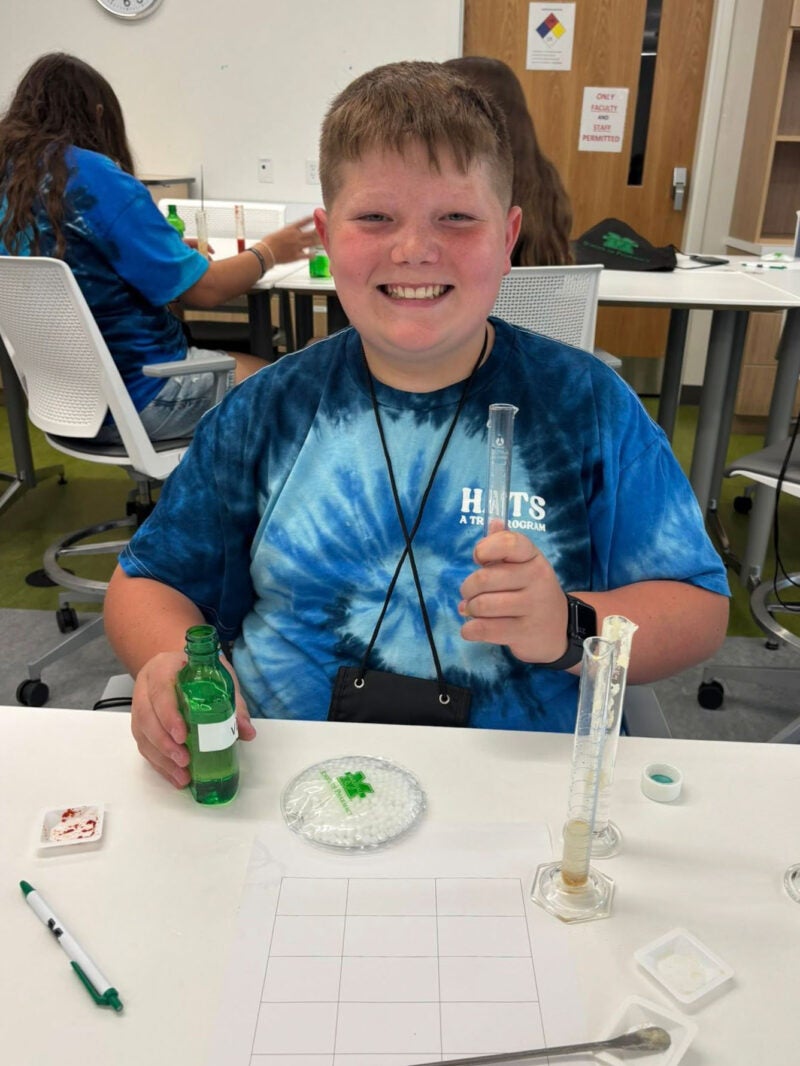 student sitting at a table performing a science experiment and smiling at the camera