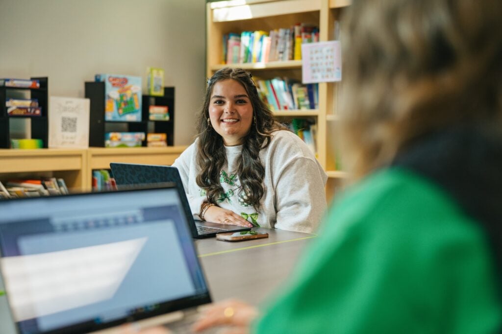 shot over the shoulder of a student working on a laptop and another student smiling at the camera