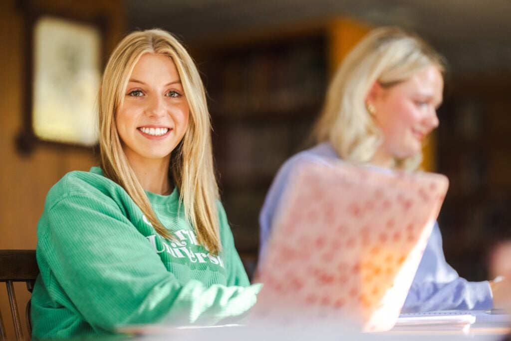 two students working in the library, one on a laptop and smiling at the camera