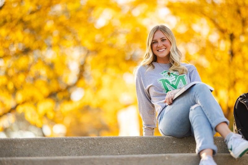 Student sitting on a bench on campus with fall leaves.